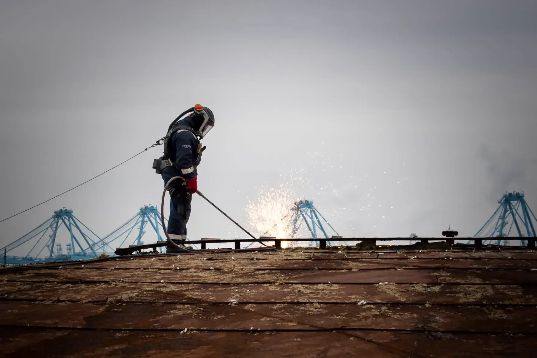 DCC-medewerker snijdt met een brander op het dak van een tank, met havenkranen op de achtergrond